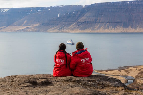Zwei Kreuzfahrt Gäste sitzen an der Küste von Island und schauen auf das Expeditionskreuzfahrtschiff im Fjord